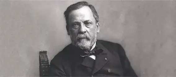 Retrato em preto e branco de um homem com barba e cabelo grisalho, vestindo fato antigo, sentado numa cadeira, olhando para a câmera.
