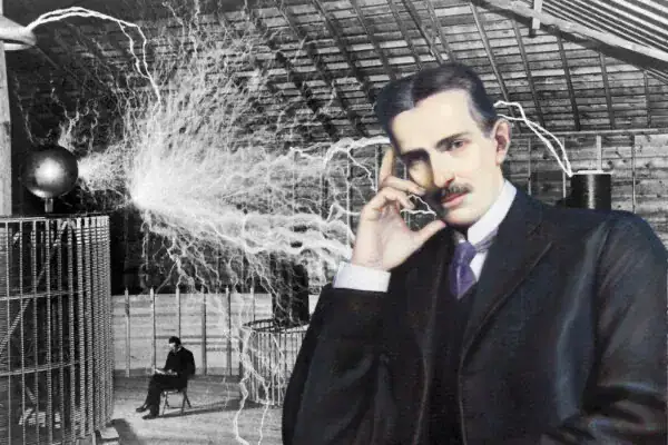 Homem com cabelo e roupa escuros, segurando o que parece ser uma bobina de Tesla, com faíscas elétricas visíveis ao fundo em ambiente industrial.