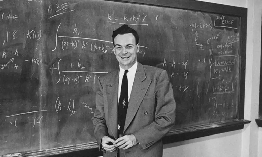 Homem sorridente de fato de treino e blazer, de pé na frente de um quadro negro com equações matemáticas escritas a giz.