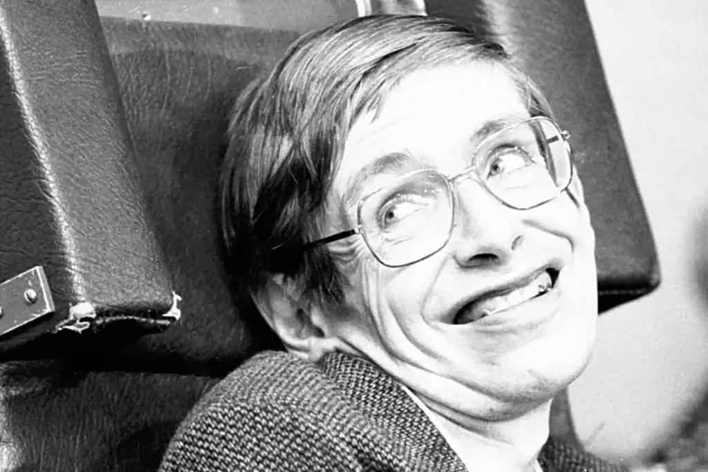 Homem com óculos de armação fina, cabelo castanho claro, sorrindo amplamente, sentado numa cadeira de escritório, com expressão alegre e descontraída.