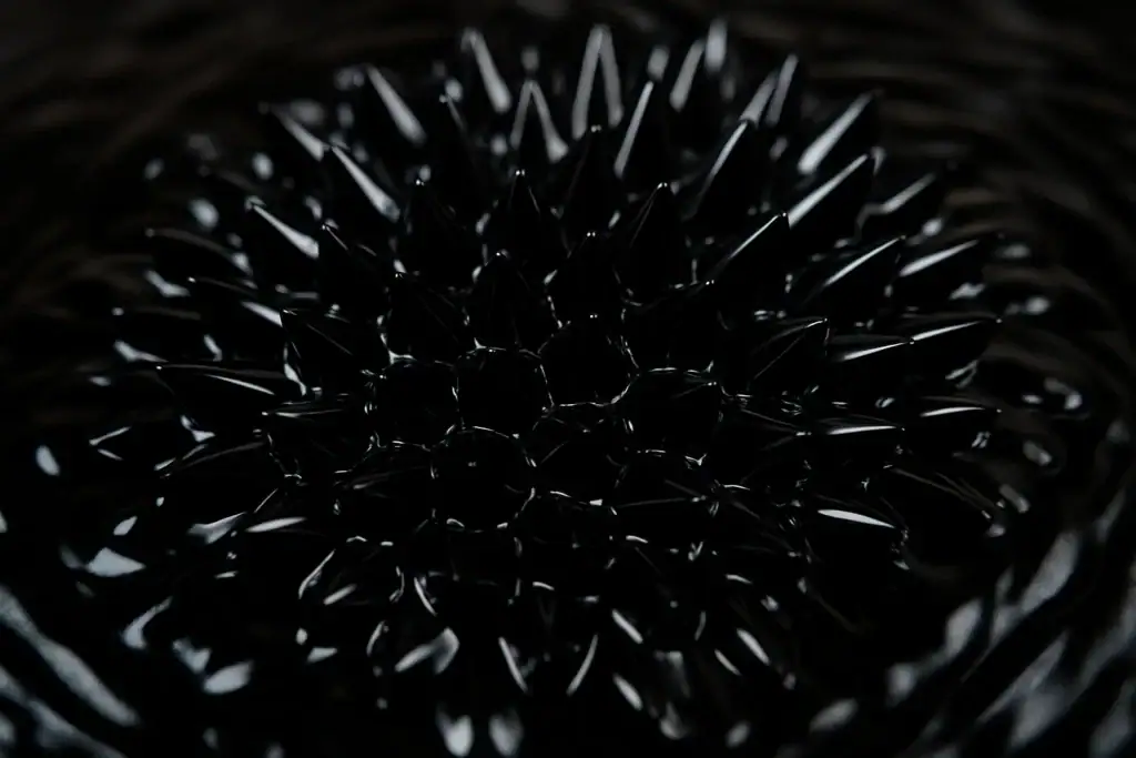 Ferrofluido preto sob efeito de campo magnético, formando padrões de spikes e linhas finas, com reflexos brilhantes na superfície.